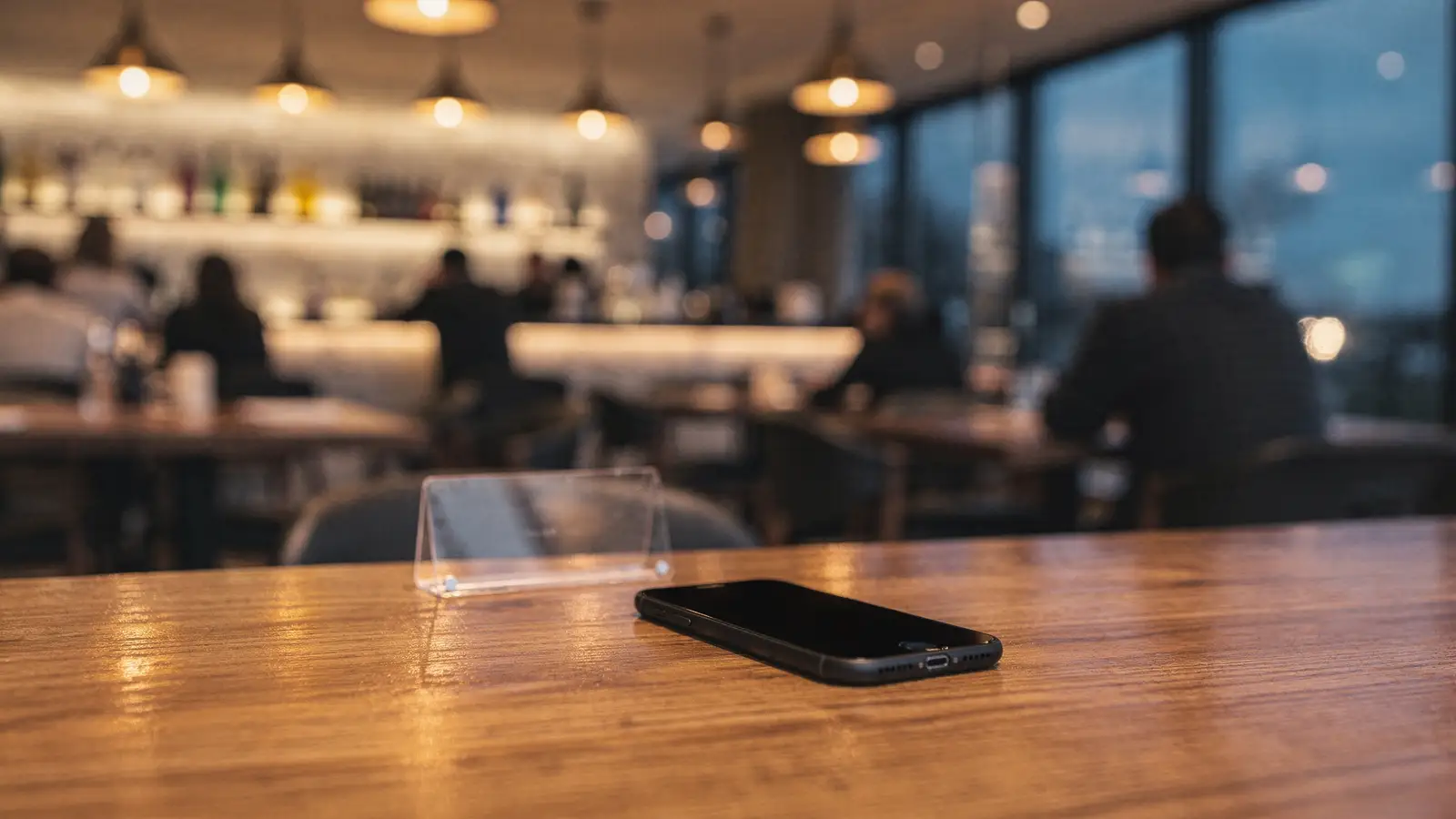 Abendliche Restaurant-Szene: Smartphone und transparenter Aufsteller auf einem Holztisch, Bar im Hintergrund